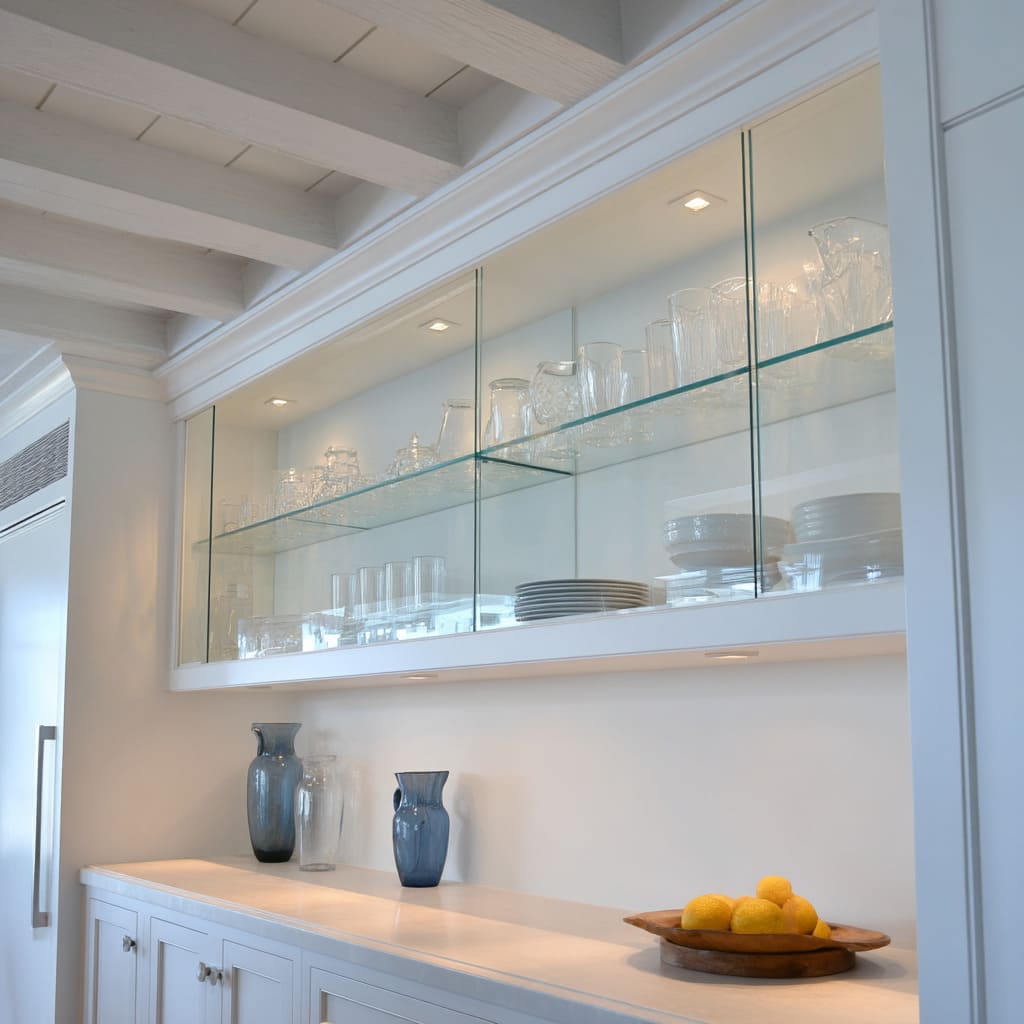 One Long Glass Upper Display Cabinet Tucked Under Ceiling Beams in a Clean Coastal White Kitchen Design One Long Glass Upper Display Cabinet Tucked Under Ceiling Beams in a Clean Coastal White Kitchen Design